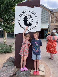 Two children smiling and standing in front of the Denver Zoo entrance sign on a sunny day.