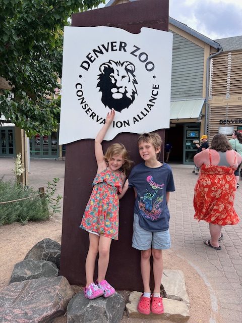 Two children smiling and standing in front of the Denver Zoo entrance sign on a sunny day.