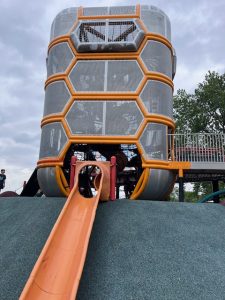 Wide-angle view of the large orange microphone-themed play structure at Paco Sánchez Park in Denver, featuring climbing nets and unique design elements under a sunny sky.