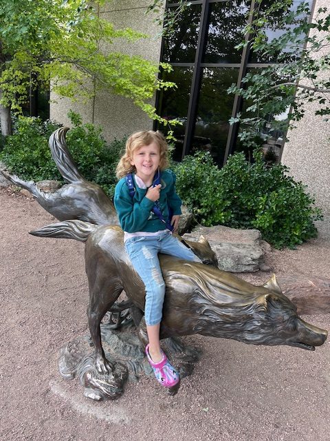 A young girl sits smiling on top of a large bronze wolf sculpture outside the Denver Museum of Nature and Science. She’s framed by the museum’s stone exterior and surrounded by greenery, capturing a playful start to a day of discovery.