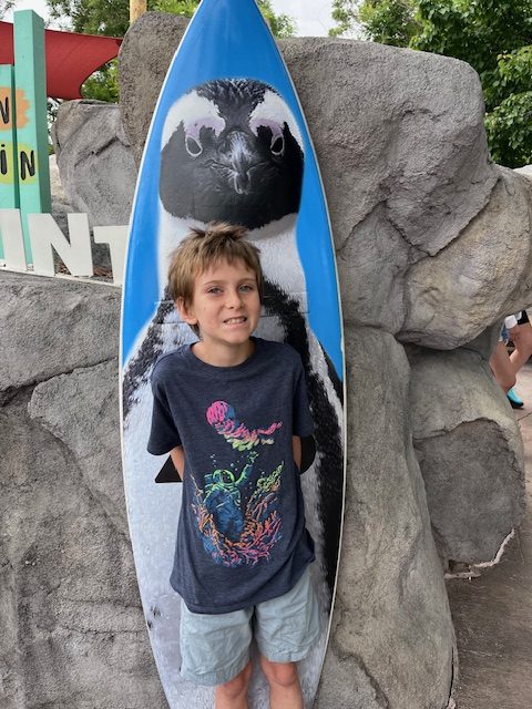 Young boy smiling while standing in front of a penguin exhibit sign at the Denver Zoo.