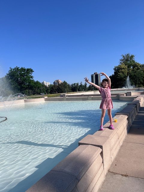 Young girl stands on the edge of a large fountain at Cheesman Park in Denver, arms raised in joyful triumph. The water sparkles behind her as she balances confidently, capturing a carefree moment of play and exploration.