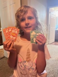 Smiling young girl holding colorful pretend money labeled ‘Ember Bucks,’ part of a homeschool reward system used to teach responsibility and encourage positive behavior.