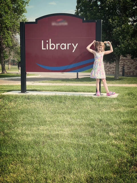A young girl in a dress standing on the grass in front of a library sign, striking a playful, silly pose with a big smile.