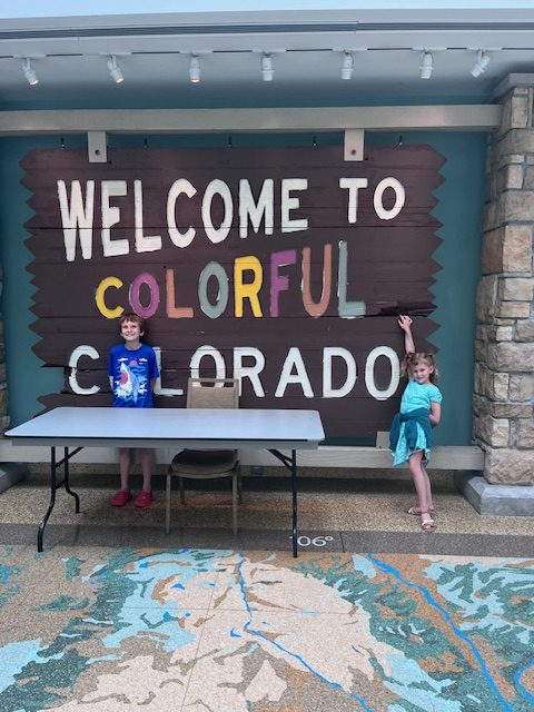 Two children stand smiling in front of a large, colorful sign that reads “Welcome to Colorful Colorado” inside the History Colorado Center. The bold, vibrant design of the sign creates a striking backdrop, capturing a playful and memorable moment from their museum visit.