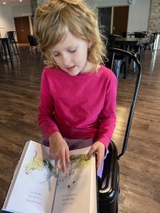 Young girl sitting cross-legged, deeply focused as she reads an All About Reading book—sunlight spilling across the pages, her face lit with quiet confidence and curiosity.