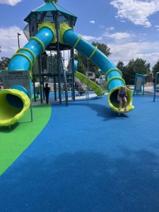 Young girl sits at the bottom of a vibrant blue and green slide at Benedict Park, smiling as she finishes her ride. The colorful play structure behind her features musical elements and climbing features, inviting kids into a world of active, sensory-rich play.