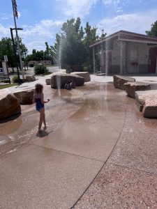 Young girl plays in the splash area at Benedict Park, running through shallow water and sprinklers with joyful energy. The creek-style splash pad offers a refreshing break from the heat, surrounded by natural elements and open space.