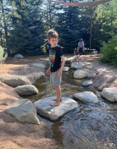 Young boy stands barefoot on a large rock in the creek-style splash area at the Mordecai Children’s Garden in the Denver Botanic Gardens. Surrounded by shallow water and smooth stones, he balances carefully, soaking in the sensory-rich nature play environment.