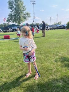 A young girl with her face painted, smiling and holding a mini golf club while standing on the grass at the library’s summer reading kickoff event.