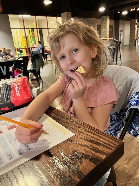 Young homeschooler sitting at a table with schoolwork, casually eating a chip—learning and snacking during a relaxed multi-grade homeschool day.
