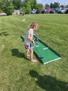 A young girl in a dress concentrating as she plays mini golf on the grass during the library’s summer reading kickoff event.