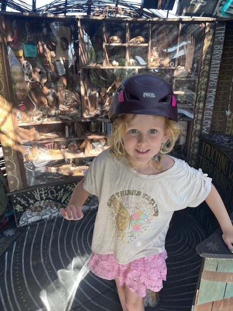 Young girl wearing a safety helmet stands at the base of the outdoor climbing structure at the Children’s Museum of Denver. She looks up with focus and anticipation, ready to begin the rope-and-platform adventure ahead.