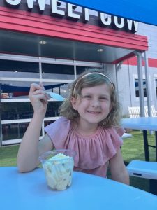Young girl enjoys a scoop of mango dairy-free ice cream at Stanley Marketplace. She smiles mid-bite, the bright orange treat glowing in the afternoon sun as she sits on the open patio, savoring a sweet moment on a warm day.
