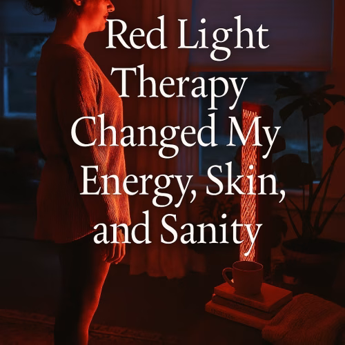 Woman standing barefoot in a softly lit living room, facing a glowing red light therapy panel. The warm red glow illuminates her face and body as she stands calmly on a patterned rug. A side table nearby holds a mug, folded blanket, and books titled “Gentle Rituals” and “Wintering.” In the background, a child plays near a window showing the cool twilight sky. Overlaid text reads: “Red Light Therapy Changed My Energy, Skin, and Sanity.”